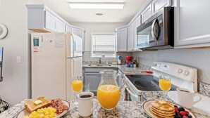 Cozinha moderna com armários brancos, geladeira, micro-ondas, fogão e espaço para café da manhã, incluindo torradas, ovos, panquecas e frutas.