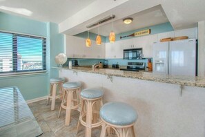 A kitchen with a bar area, bar stools, a refrigerator, and a window with blinds. - Beach Club II 9E - Amazing Oceanfront Views (North Myrtle Beach)