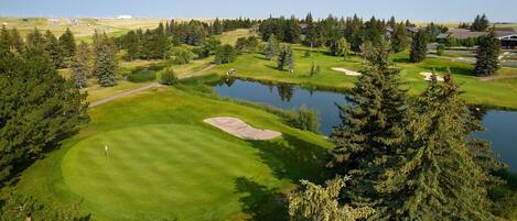 Un campo de golf con un estanque, árboles y edificios al fondo.