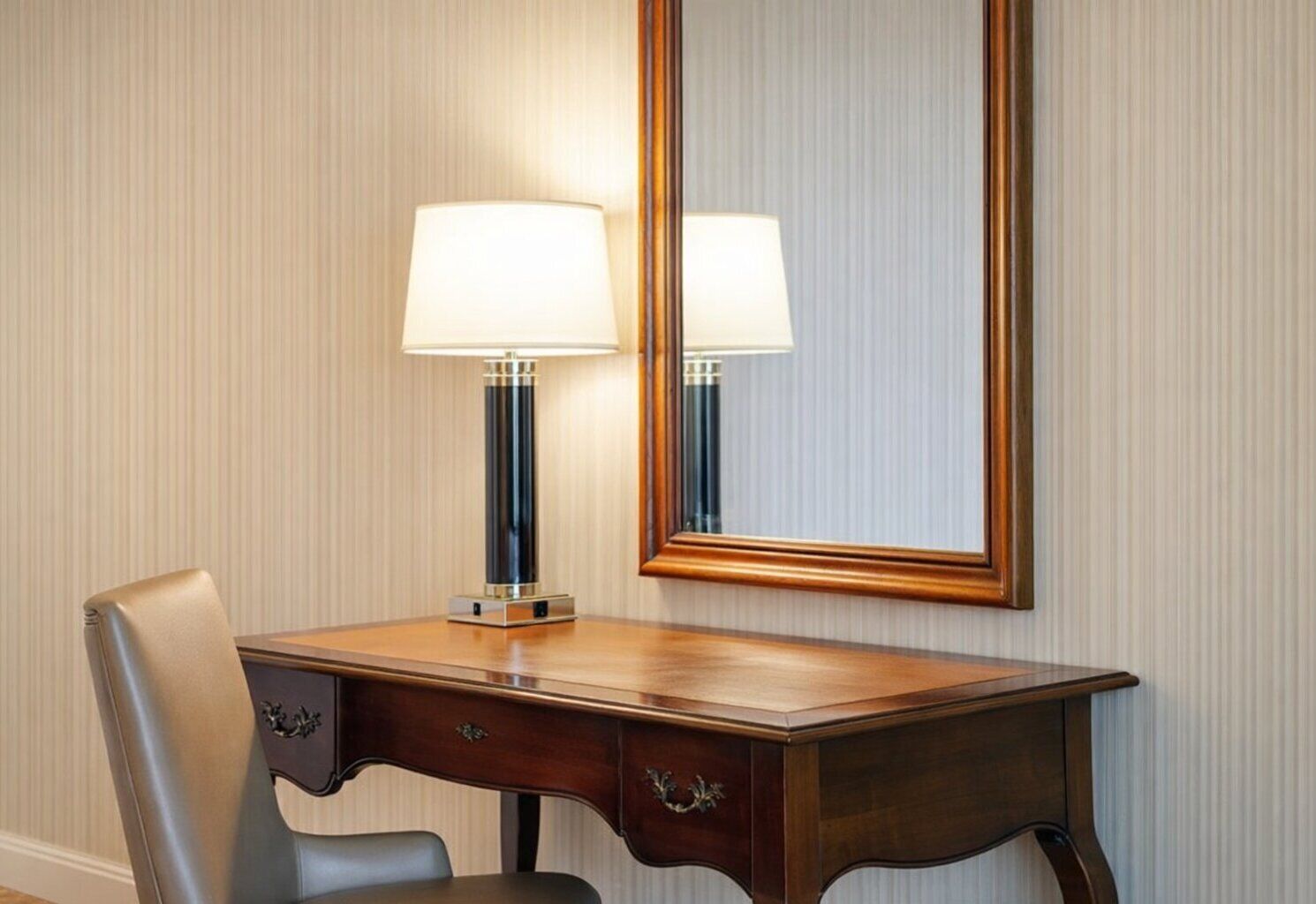 A hotel room with a wooden desk, two lamps, a mirror, and a chair.