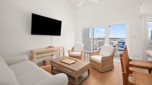 A living room with a flat-screen TV, a sofa, a coffee table, wicker chairs, and a balcony with a view. - Three Bedroom Bay View Townhome in Fenwick Island (Fenwick Island)