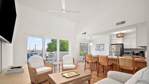 A modern living room with a sofa, wicker chairs, a wooden coffee table, and a dining area with a bar. - Three Bedroom Bay View Townhome in Fenwick Island (Fenwick Island)