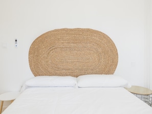 Ein Bett mit weißer Bettwäsche, ein runder, gewobener Kopfteil, ein kleiner runder Tisch mit Holzbeschichtung und eine weiße Wand mit Steckdose und Lichtschalter.