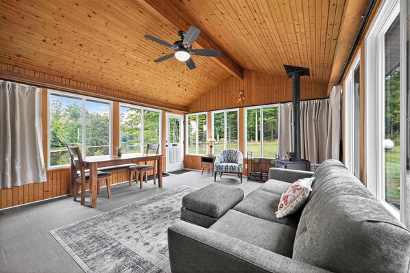 A spacious room with a wooden ceiling, large windows, a ceiling fan, a sofa, a dining table, and a chair.