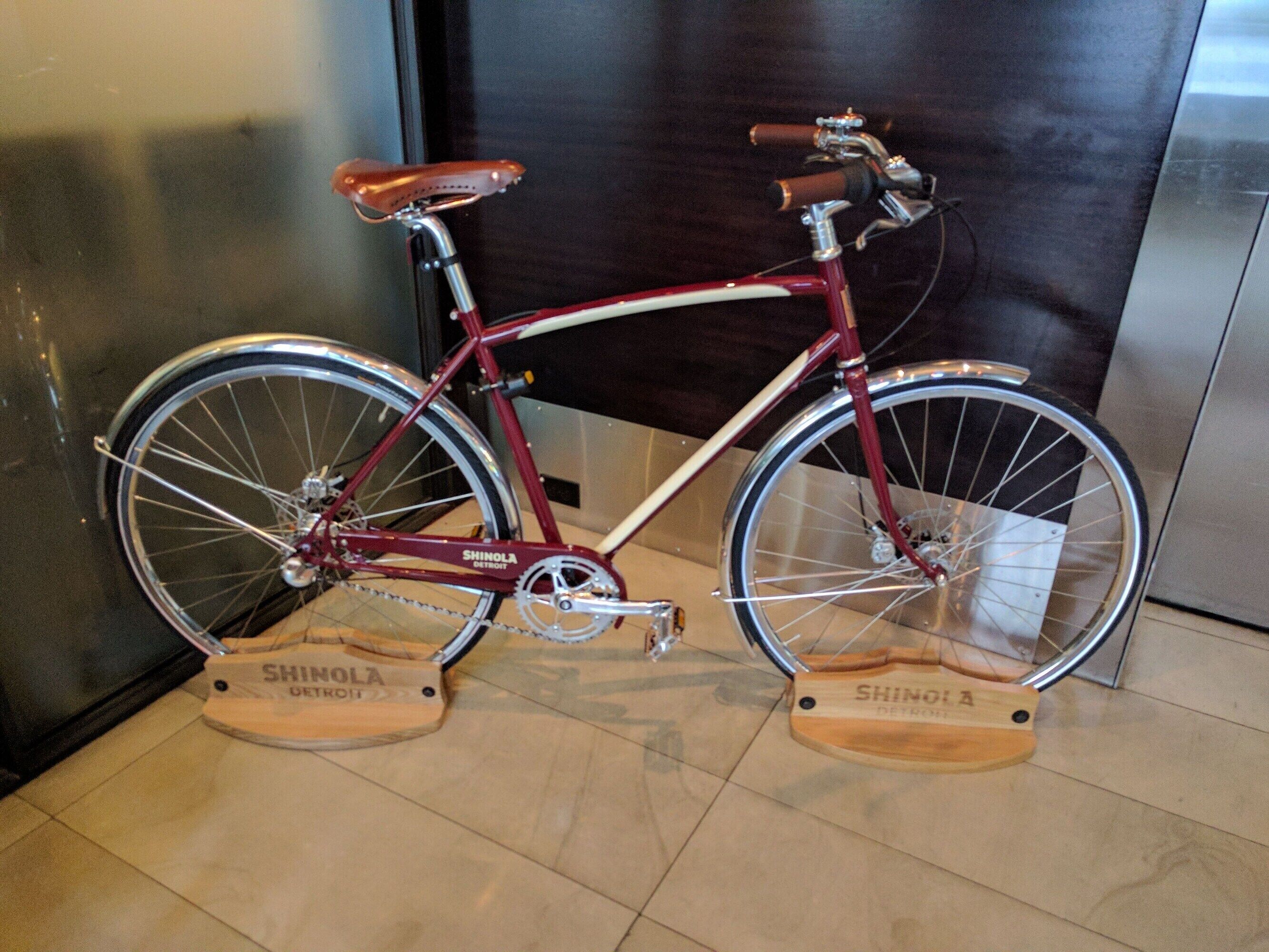 Uma bicicleta vermelha com pedais de madeira e um assento marrom, posicionada sobre um piso de azulejos com dois blocos de madeira rotulados 'SHINOLA DETROIT' embaixo.