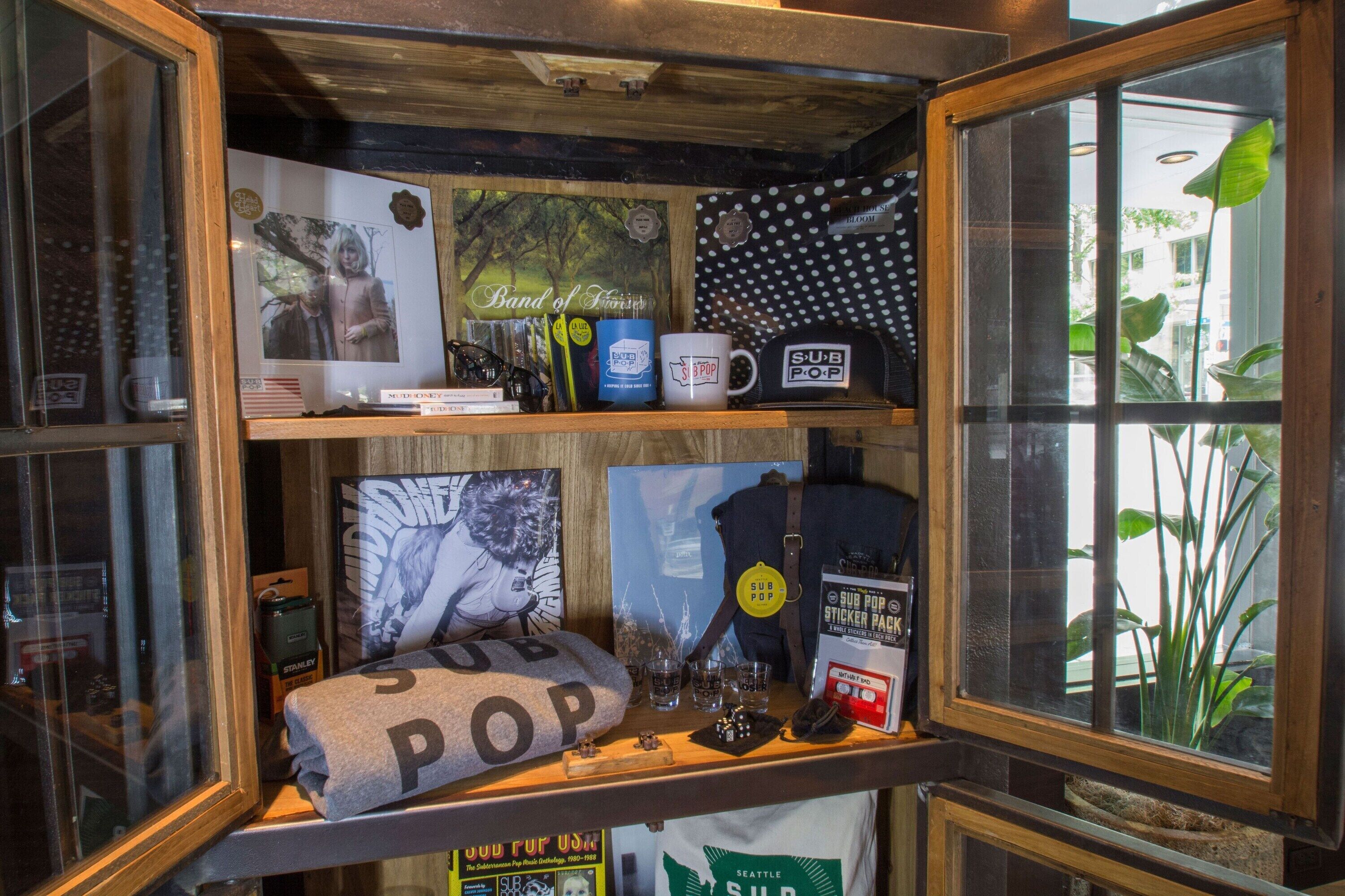 A display case with various items including a 'Band of Brothers' poster, a polka dot bag, a 'Sub Pop' pillow, a mug, and a 'Sub Pop' sticker pack.