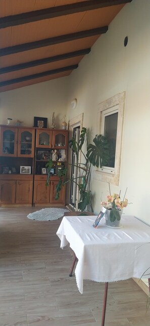 Une pièce avec un plafond en bois, une table blanche ornée d’un vase de fleurs, un meuble en bois aux portes vitrées et une fenêtre avec un cadre.