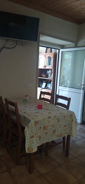 Un coin repas avec une table recouverte d’une nappe à carreaux, des chaises en bois, un téléviseur fixé au mur et une porte-fenêtre coulissante donnant sur un espace cuisine.