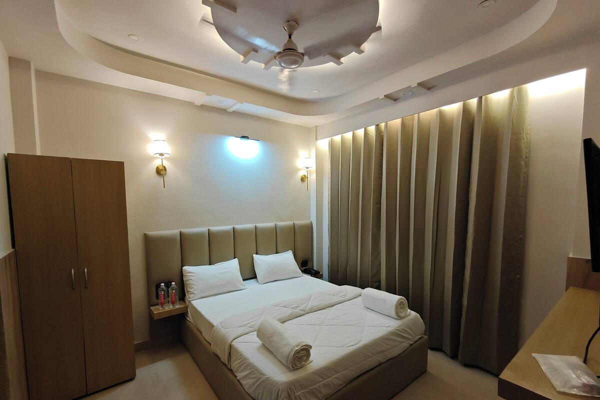 A hotel room with a bed, wardrobe, bedside table, wall sconces, curtains, and a ceiling fan.