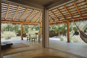 Una habitación con techo de paja, ventanas amplias, una mesa y un banco.