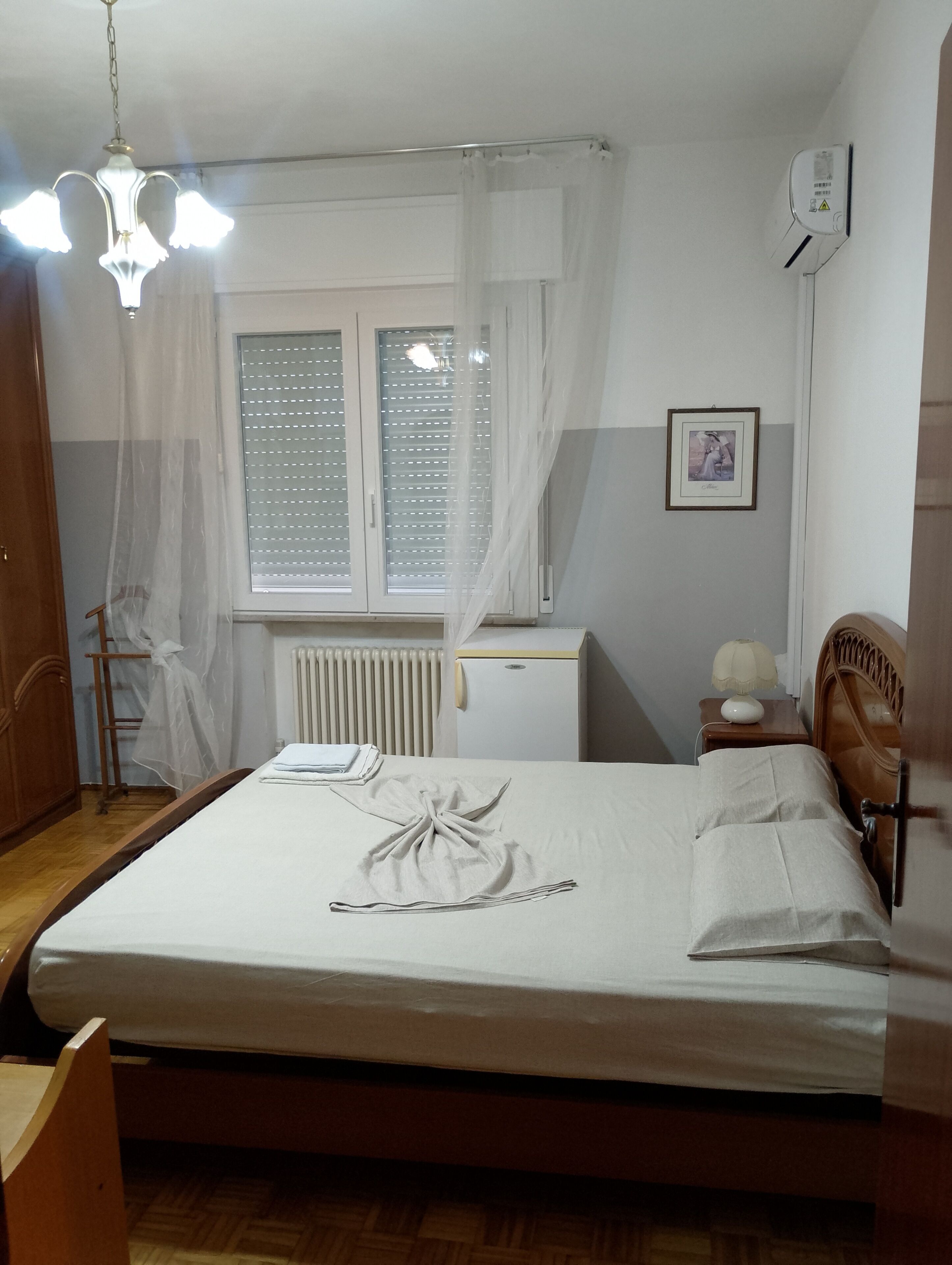 Una camera da letto con un letto, un termosifone, una finestra con persiane, un comodino con una lampada e un quadro appeso al muro.