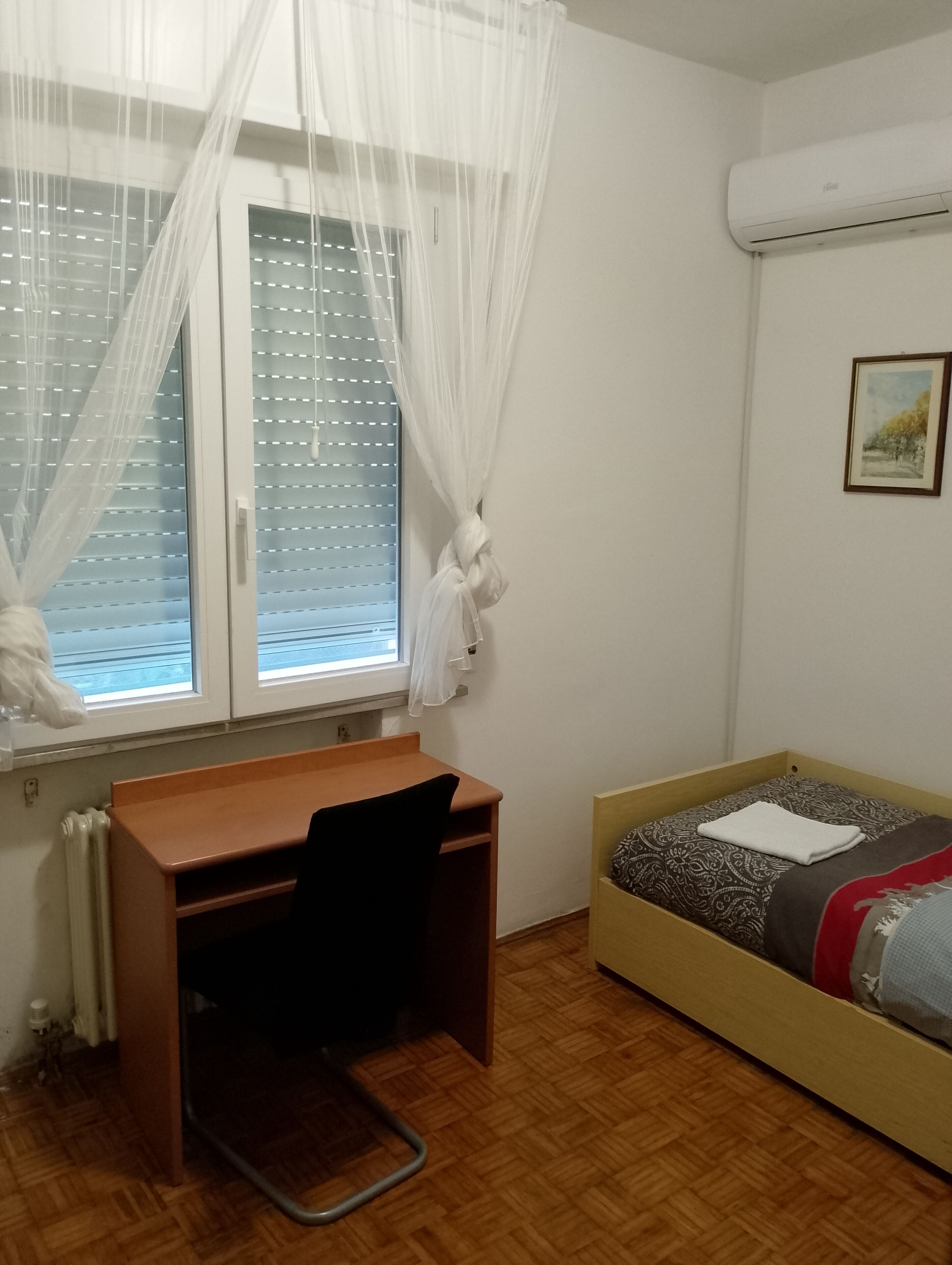 Un dormitorio con un escritorio de madera, una cama con ropa de cama estampada, una ventana con cortinas translúcidas y un equipo de aire acondicionado en la pared.