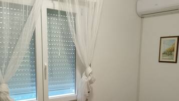 Un dormitorio con un escritorio de madera, una cama con ropa de cama estampada, una ventana con cortinas translúcidas y un equipo de aire acondicionado en la pared.
