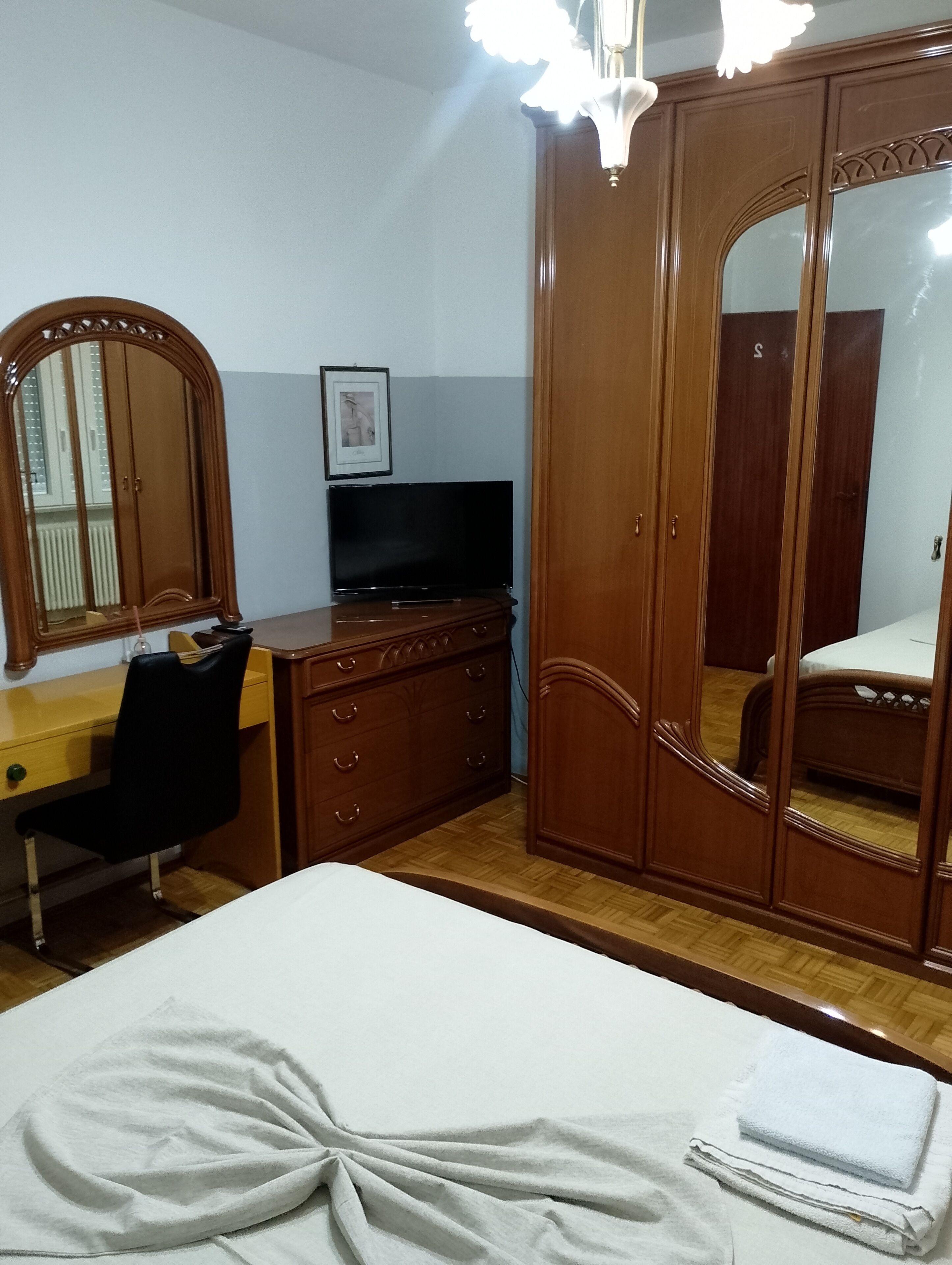Ein Schlafzimmer mit einem Bett, einem Schminktisch, einem Schreibtisch mit Stuhl, einem Fernseher und einem Kleiderschrank mit Spiegel.