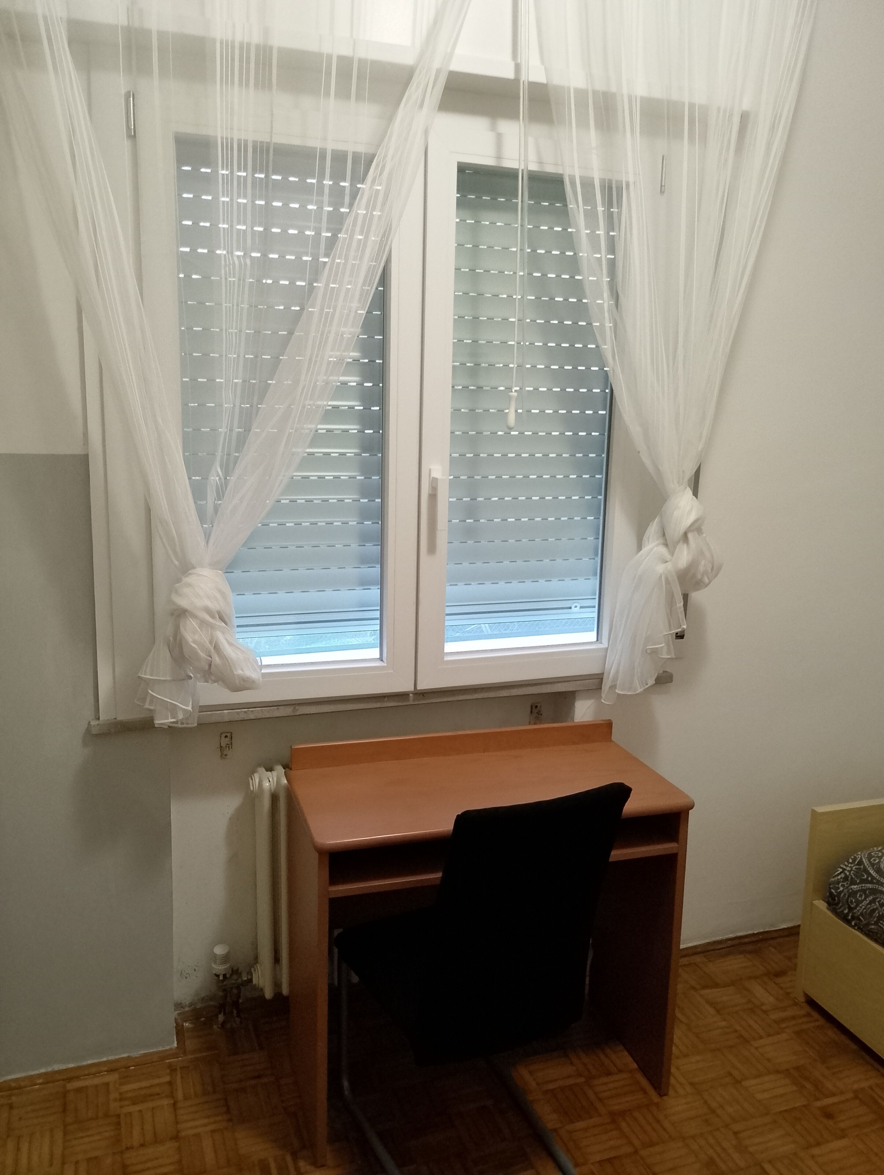 Et værelse med et træskrivebord, en stol, en radiator og et vindue med lette gardiner.