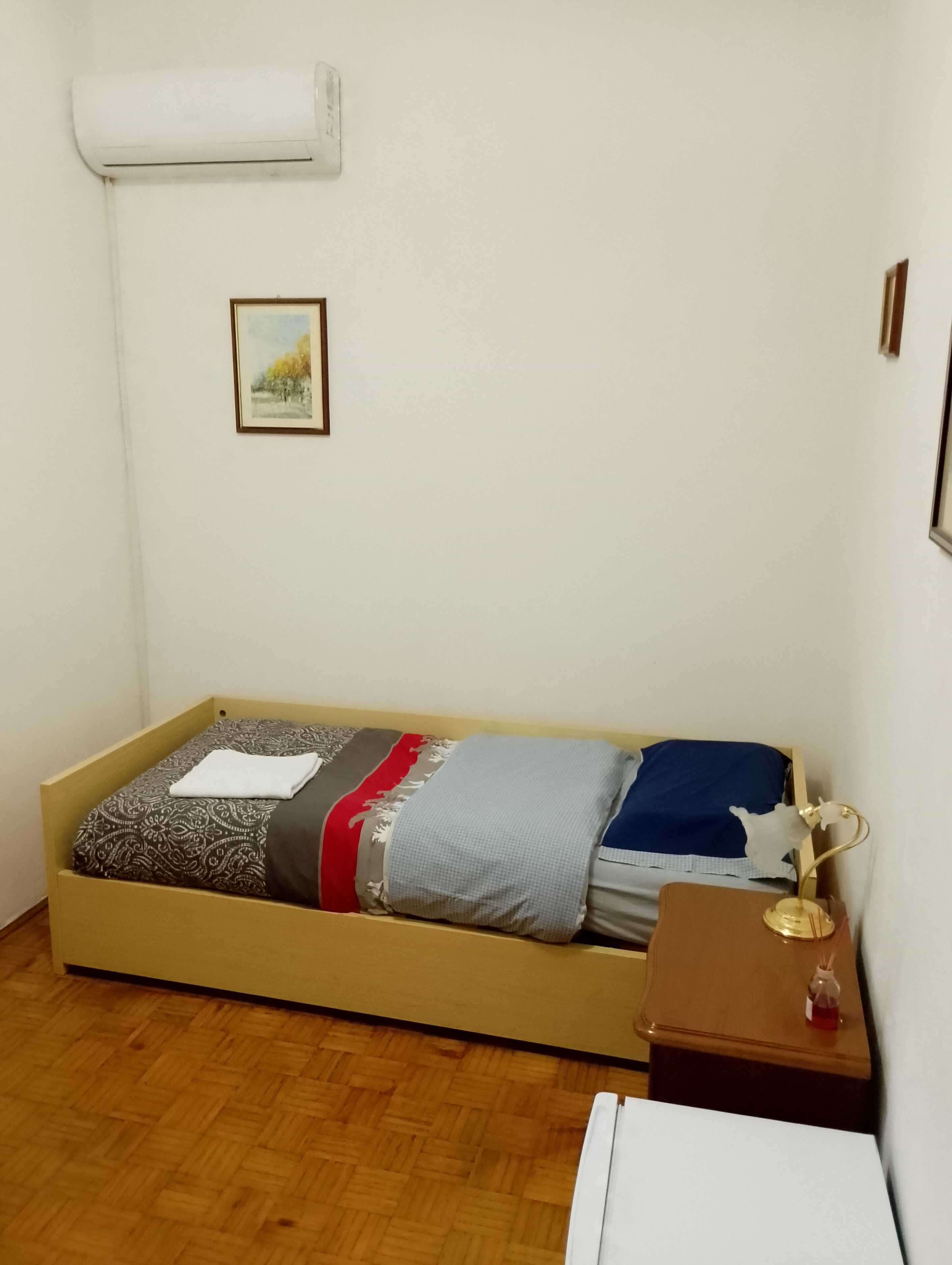 A single bed with a patterned bedspread, a bedside table with a lamp, a picture on the wall, and a white air conditioning unit.