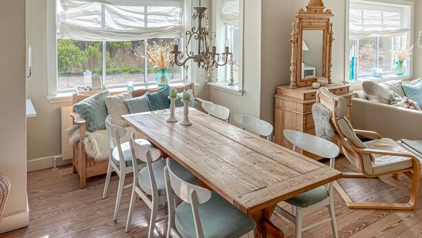 Un comedor con una mesa de madera, sillas blancas, una araña y un ventanal con vistas a la vegetación.