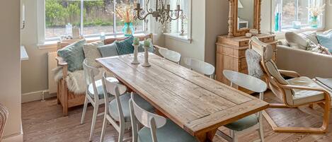 Un comedor con una mesa de madera, sillas blancas, una araña y un ventanal con vistas a la vegetación.