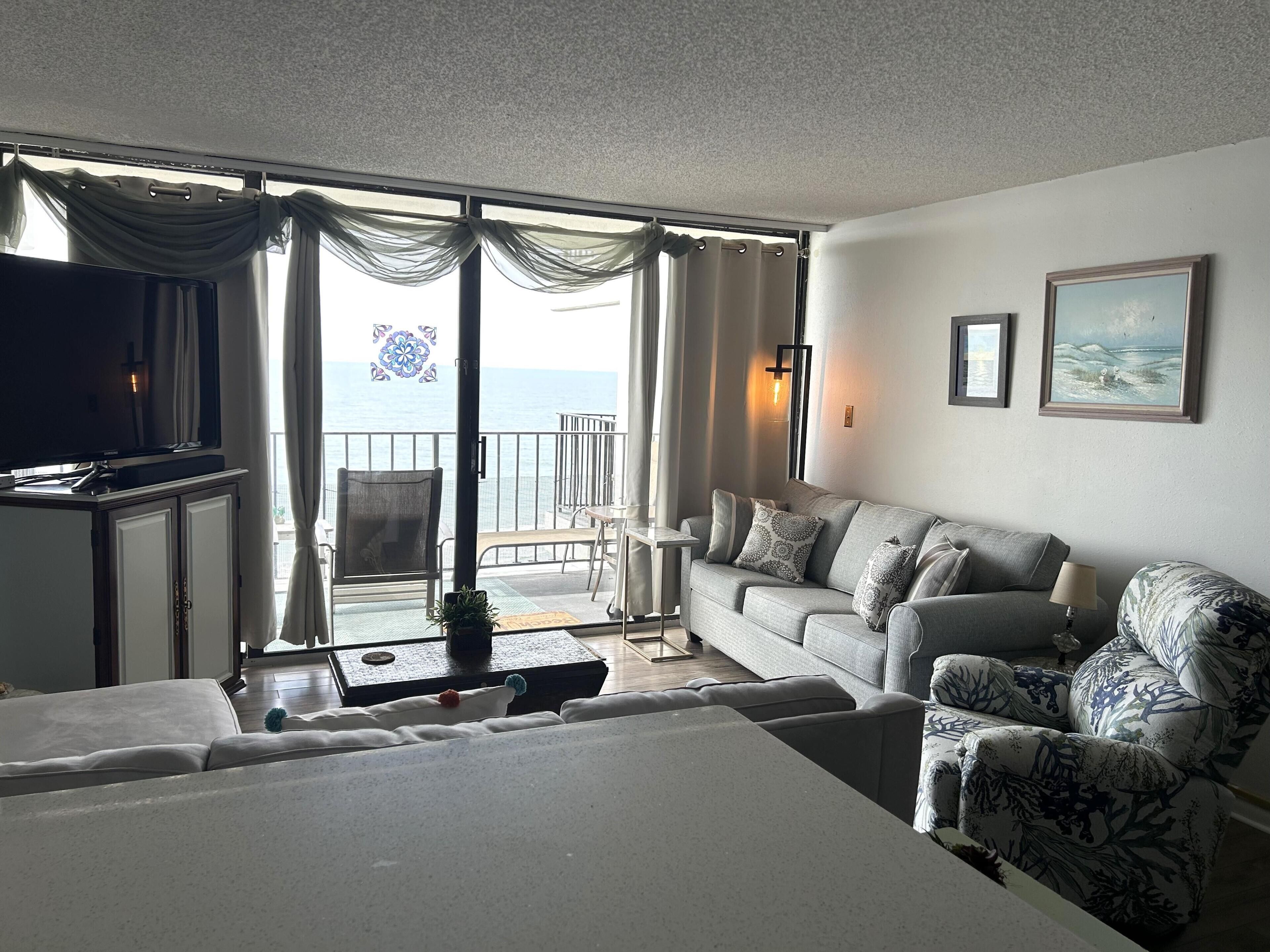 Een woonkamer met een bank, fauteuils, een televisie en uitzicht op zee door grote ramen.