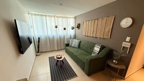 Uma sala de estar com um sofá verde, uma televisão de tela plana, um relógio e um vaso de planta.