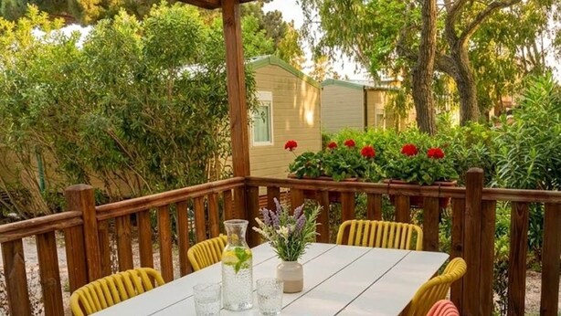 A wooden deck with a white table and yellow and orange chairs, surrounded by greenery and a small building. - Mobile home with 4 bedrooms for 8 people (Sainte-Marie-la-Mer)