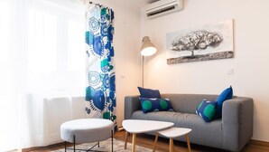 Una sala de estar con un sofá gris, cojines azules y verdes, un otoman redondo, una mesita blanca y un cuadro de un árbol.