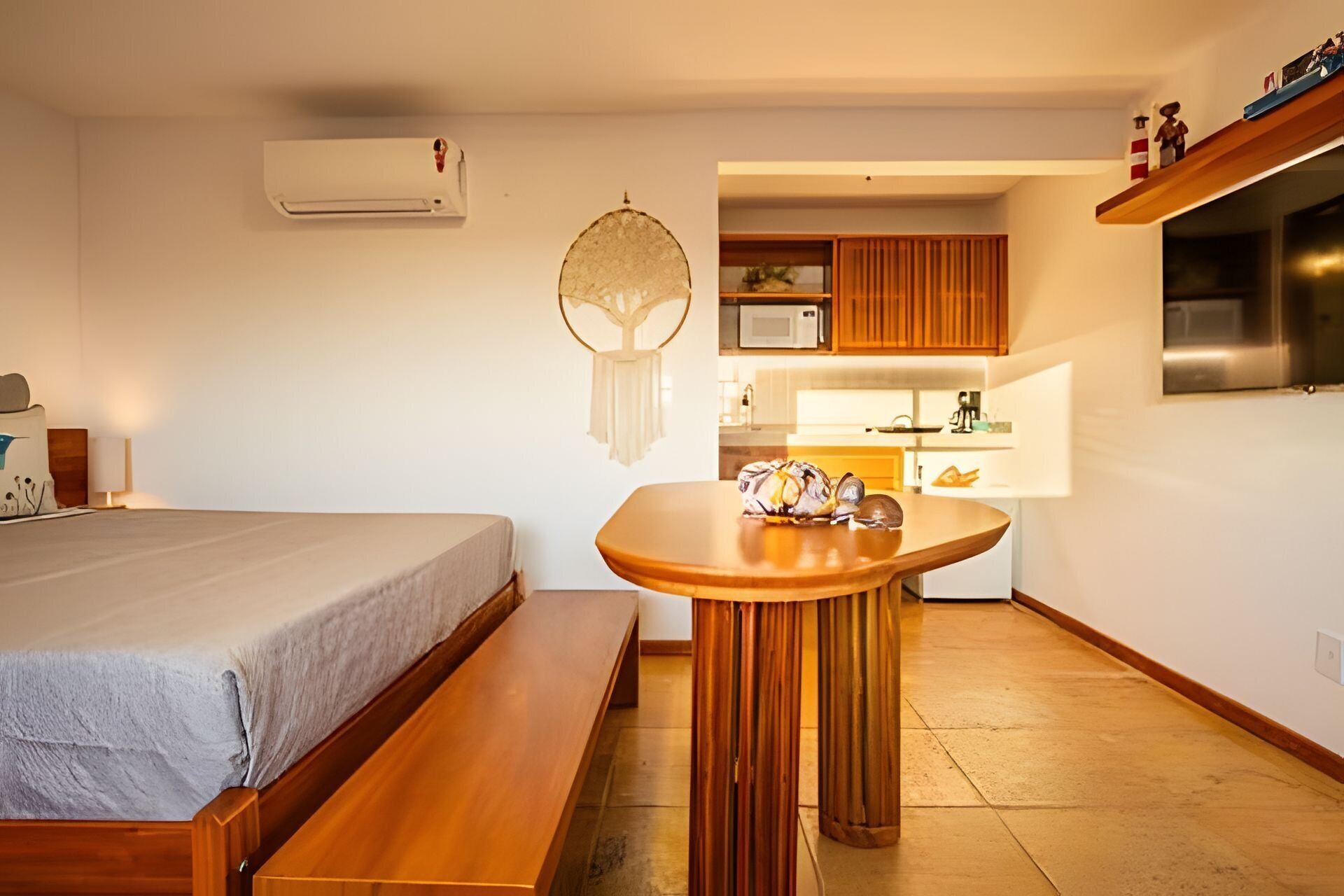 Une pièce de taille modeste comprenant un lit, une petite table en bois, un climatiseur fixé au mur, un coin cuisine avec des placards en bois et un four à micro-ondes.