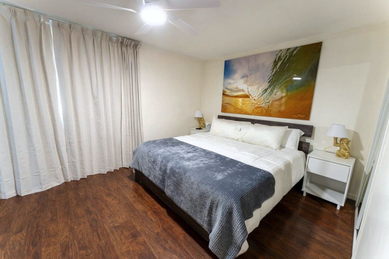 A hotel room with a bed, a nightstand, a lamp, a ceiling fan, and a painting on the wall.