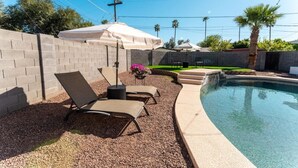 Un patio trasero con piscina, dos sillones de descanso, una mesita y un área de patio techada.