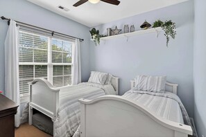 A bedroom with two beds, a window with blinds, a ceiling fan, and a shelf with decorative items. - Tuna Luna - Coastal Getaway Essentials Included (Carolina Beach)