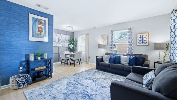 A modern living room with a blue accent wall, a grey sofa, a dining area with a table and chairs, and a kitchen in the background. - Ice Bath, Fire Pit & Outdoor Bar · 3BR House · Uptown Charlotte (Charlotte)