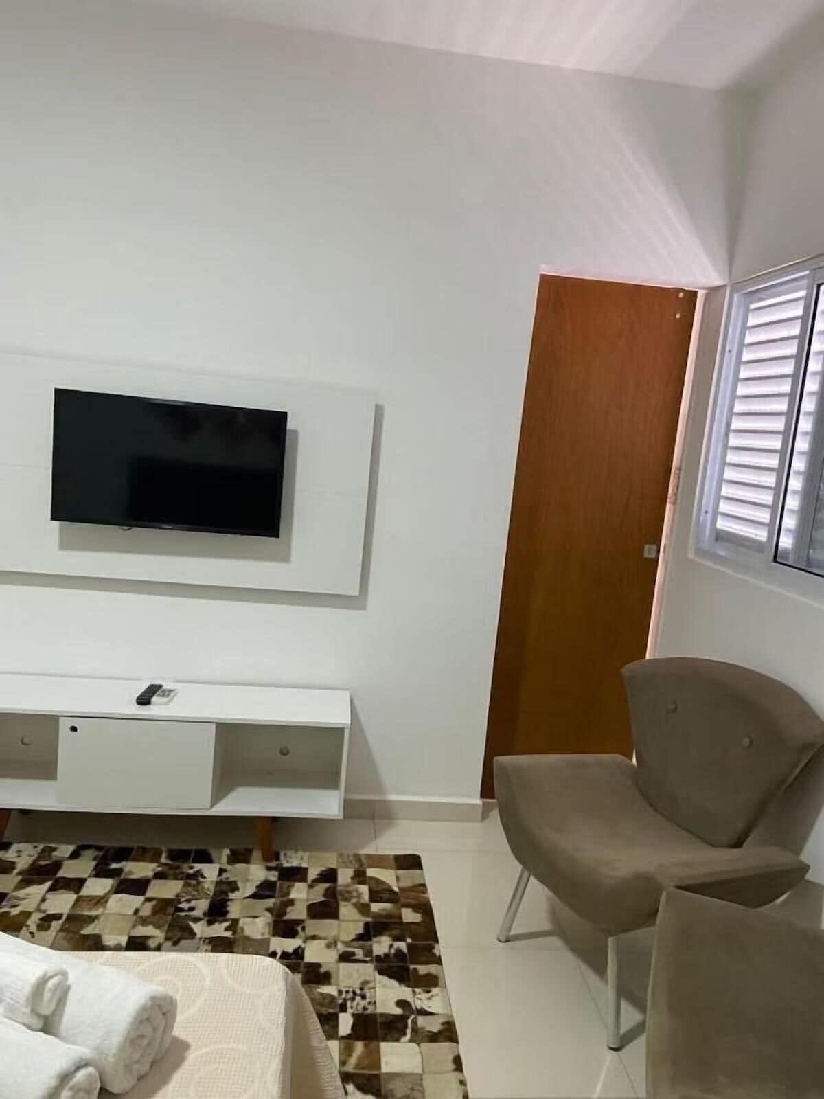 Un dormitorio con televisión montada en la pared, una puerta de madera, una ventana con persianas y una alfombra estampada.