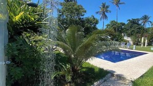 A pool with a clear blue sky, surrounded by lush greenery and palm trees. - Pousada Villa Novo Tempo (Guarujá)