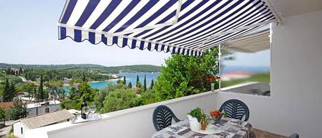 A balcony with a striped awning, a table with a tablecloth, and a view of a coastal landscape.