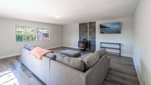 Een moderne woonkamer met een grijze bank, een open haard, een aan de muur bevestigde televisie en een raam met uitzicht op groen.