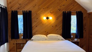 Ein Schlafzimmer mit schräg verlaufender Holzbalkendecke, einem Bett mit weißer Bettwäsche, zwei Nachttischen und einer Wandleuchte.