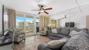 Ein modernes Wohnzimmer mit einer Couch, Sesseln, einem Fernseher, Deckenventilatoren und großen Fenstern mit Blick auf die Stadt.