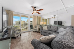 A modern living room with a sofa, armchairs, a TV, ceiling fans, and large windows offering a city view. - Dunes of Panama 2BR / 2BA Beachfront Condo C401 (Panama City Beach)