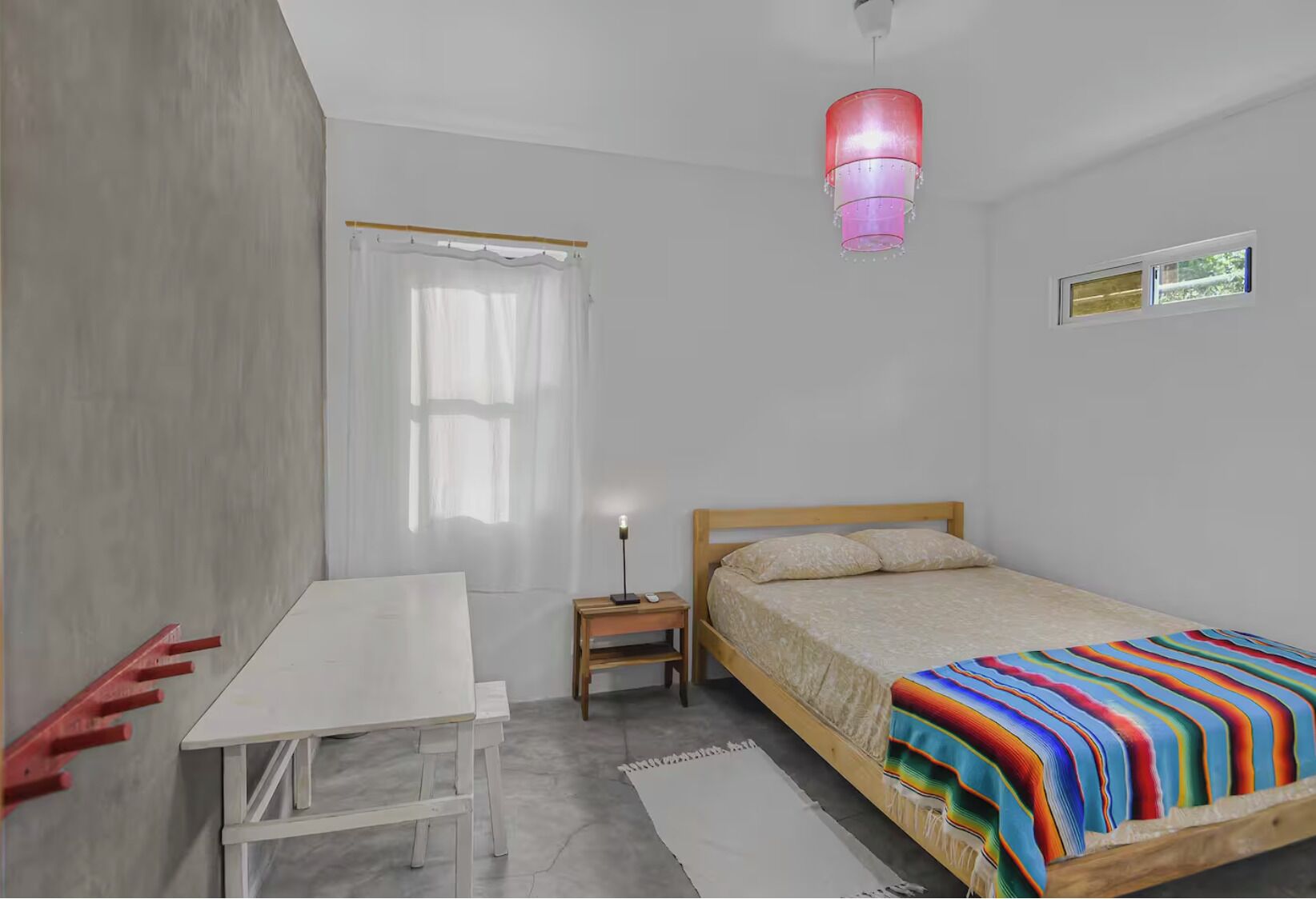 A bedroom with a bed, a desk, a chair, a nightstand, a lamp, a window, and a hanging light fixture.