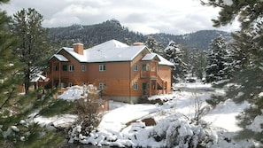 Un chalet en bois à plusieurs étages, dont le toit est recouvert de neige, entouré de pins et avec une chaîne de montagnes en arrière-plan.