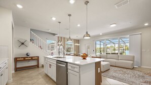 A modern kitchen with a central island, white cabinetry, and a dining area with a sofa and large windows. - 5 BD/ 4.5 BA/ Windsor Cay (16100 PF) (Clermont)