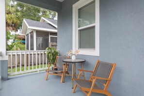 A porch with two wooden chairs and a small round table, a white vase with flowers, and a view of a house in the background. - Downtown Bungalow - 2BR Walk to Riverwalk & Armature Works 9 Min to Bucs Stadium (Tampa)