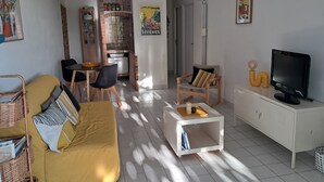 A living room with a yellow sofa, a white coffee table, a TV on a white cabinet, and a kitchen in the background.