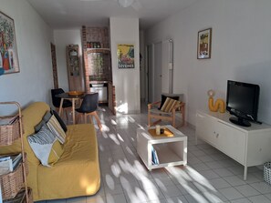Una sala de estar con un sofá amarillo, una mesita de centro blanca, un televisor sobre un mueble blanco, y una cocina al fondo.