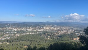 A panoramic view of a city with a dense urban area, surrounded by forested hills and distant mountains under a clear sky.