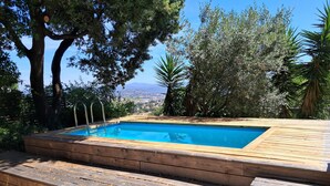 A rectangular swimming pool with a wooden deck, surrounded by trees and a view of a distant landscape. - Spacious, air-conditioned one-bedroom apartment near the sea, with a terrace, a pool, and a view of Hyères. (Hyères)
