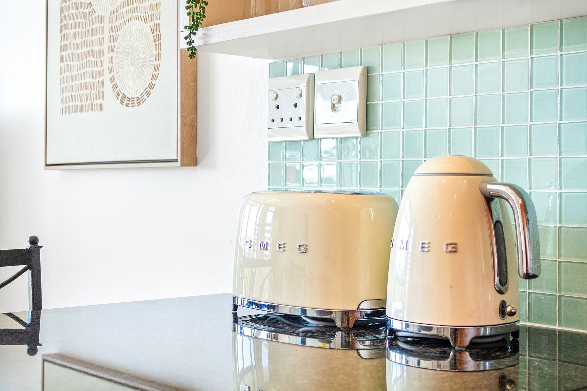 A kitchen with a coffee maker and a kettle on the counter, a framed picture on the wall, and a switchboard.