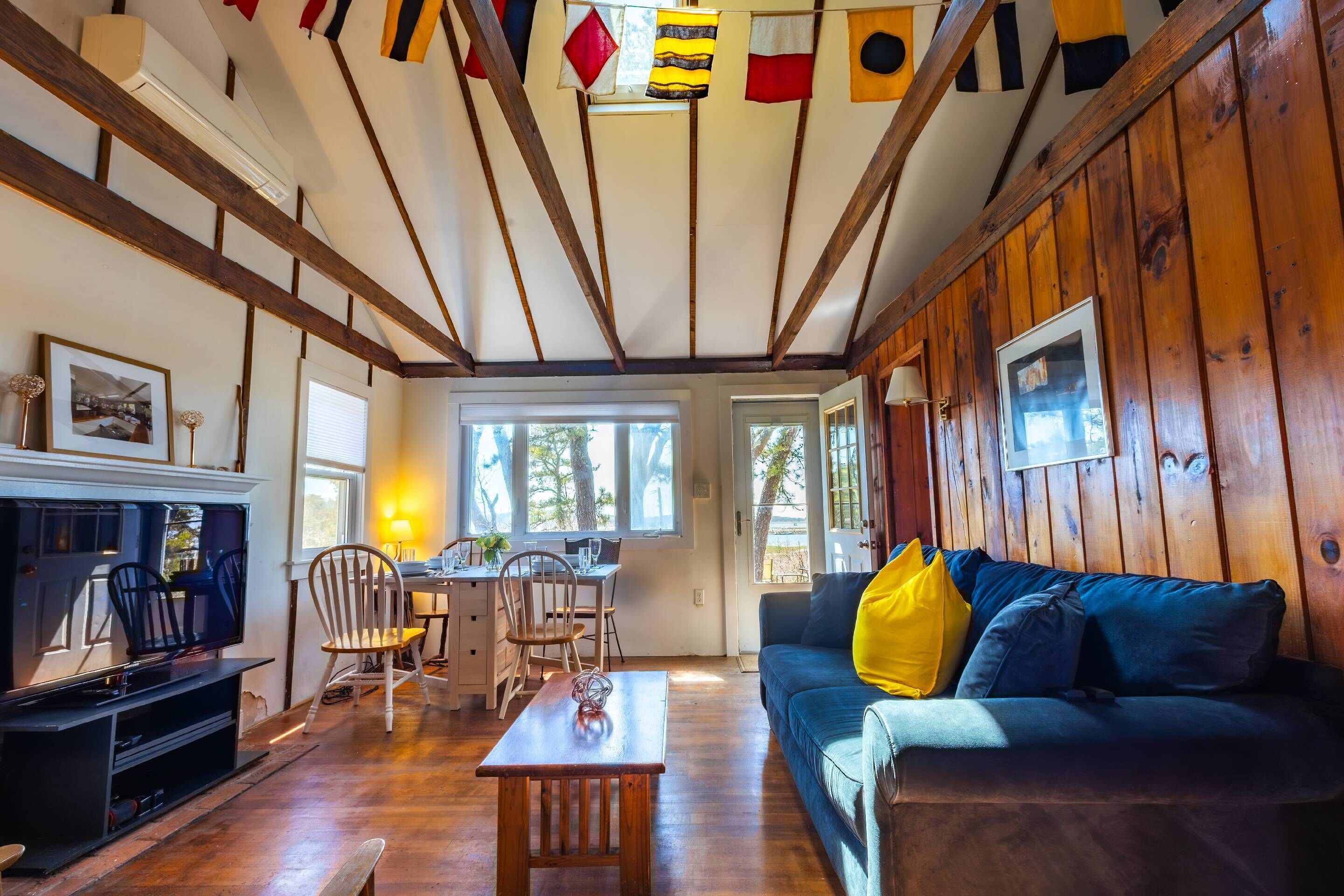 Un salon chaleureux avec un plafond en bois, un canapé bleu agrémenté de coussins jaunes, un téléviseur et un coin repas avec des chaises et une table.