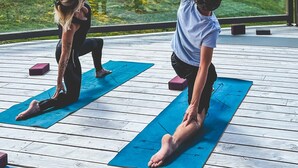 Dos personas practicando yoga sobre esterillas azules al aire libre, con una terraza de madera y edificios al fondo.