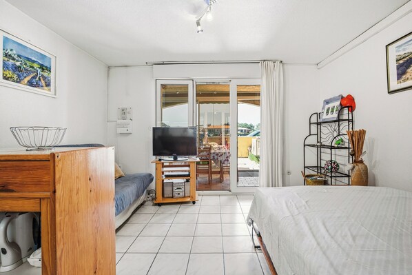 A hotel room with a bed, a TV on a wooden stand, a desk with a basket, and a view of the outside. - Le Patio d'Agde - Terrasse - Plage (Agde)